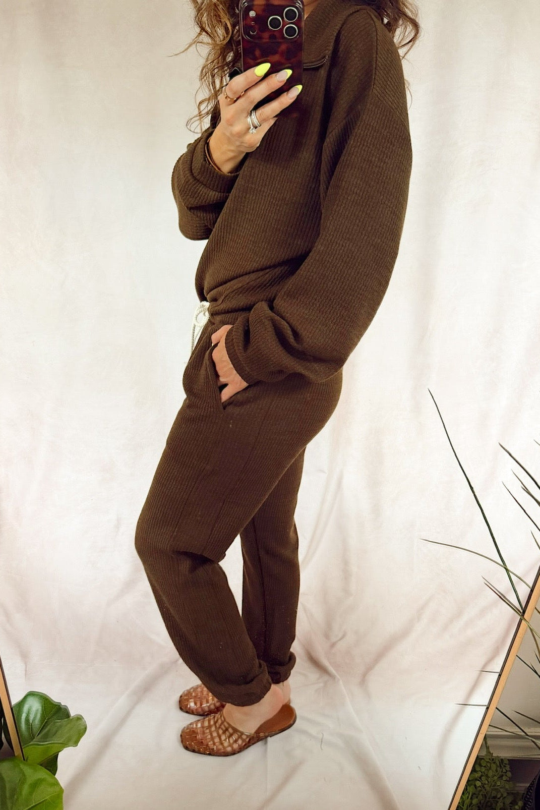 A person takes a mirror selfie wearing a matching brown knit lounge set consisting of a long-sleeve top and jogger pants, paired with brown woven flat shoes, set against a draped white fabric backdrop with indoor plants.