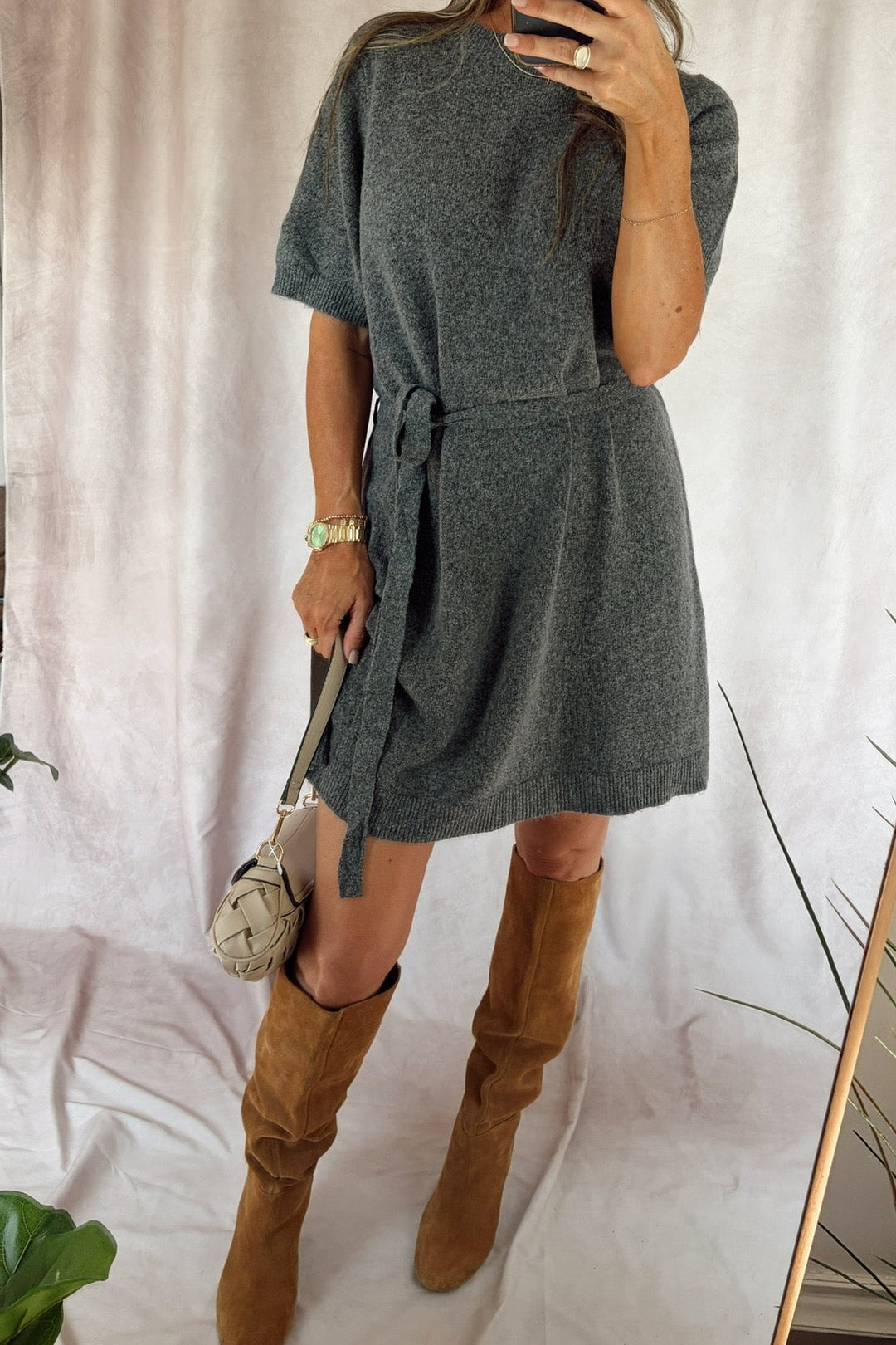 A woman takes a mirror selfie wearing a short-sleeve gray knit dress with a tie waist and brown knee-high boots, holding a small beige handbag against a draped white fabric backdrop.