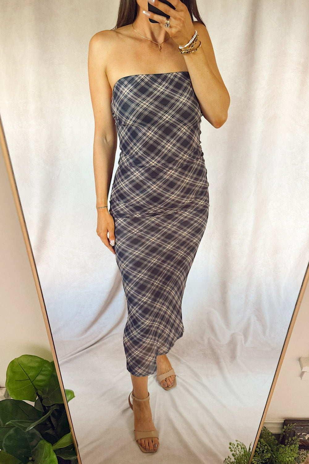 A woman takes a mirror selfie wearing a strapless plaid midi dress in dark gray and white, paired with beige heeled sandals, set against a white draped backdrop.
