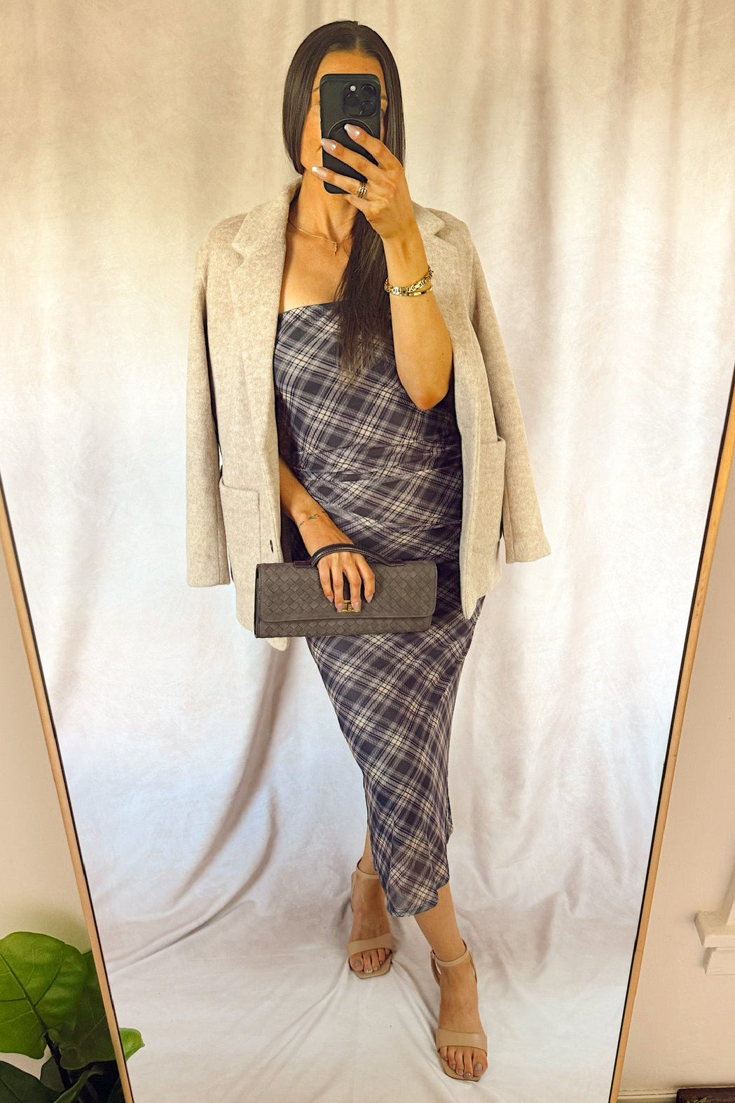 A woman takes a mirror selfie showing an outfit consisting of a light beige blazer, a blue and gray plaid strapless dress, and beige heeled sandals, holding a dark gray clutch.