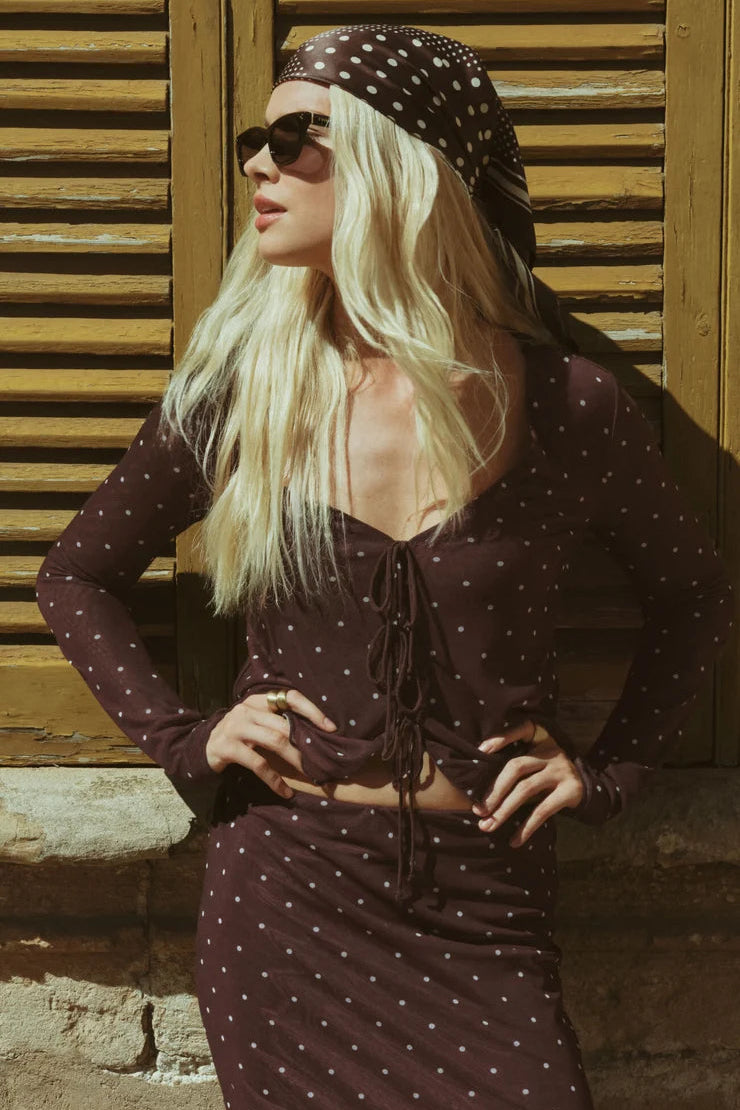 A woman wearing a dark brown long-sleeve mesh top with white polka dots, a matching polka dot headscarf, and sunglasses, posing in front of a rustic wooden shutter background.