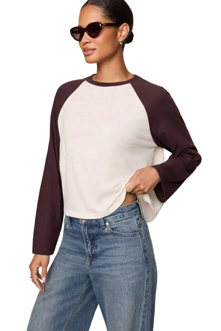 A woman modeling a long-sleeve raglan crop top with an off-white body and dark plum sleeves, paired with blue jeans against a plain beige studio background.
