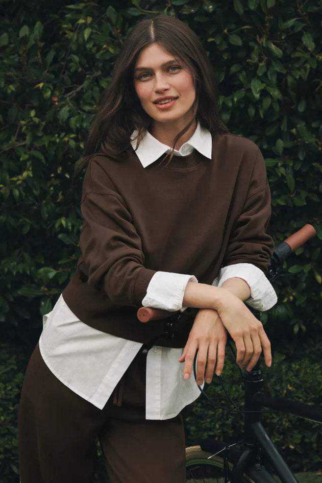 A woman wearing a brown long-sleeve top and matching brown trousers over a white collared shirt, leaning on a bicycle in front of a green hedge.