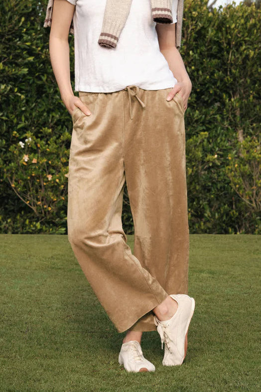 A woman stands outdoors on a grassy lawn in front of a green hedge, wearing a white short-sleeve top, beige faux suede light sand wide-leg drawstring pants, a light beige scarf, and white sneakers.