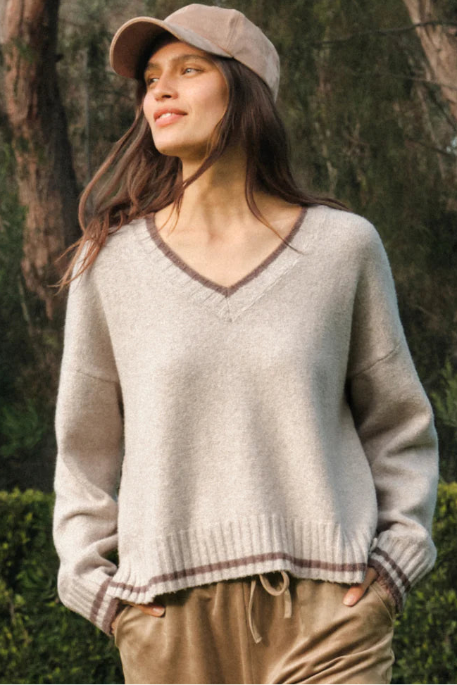 A woman wearing a beige v-neck knit sweater with brown trim, tan pants, and a matching beige cap, standing outdoors in front of trees and hedges.