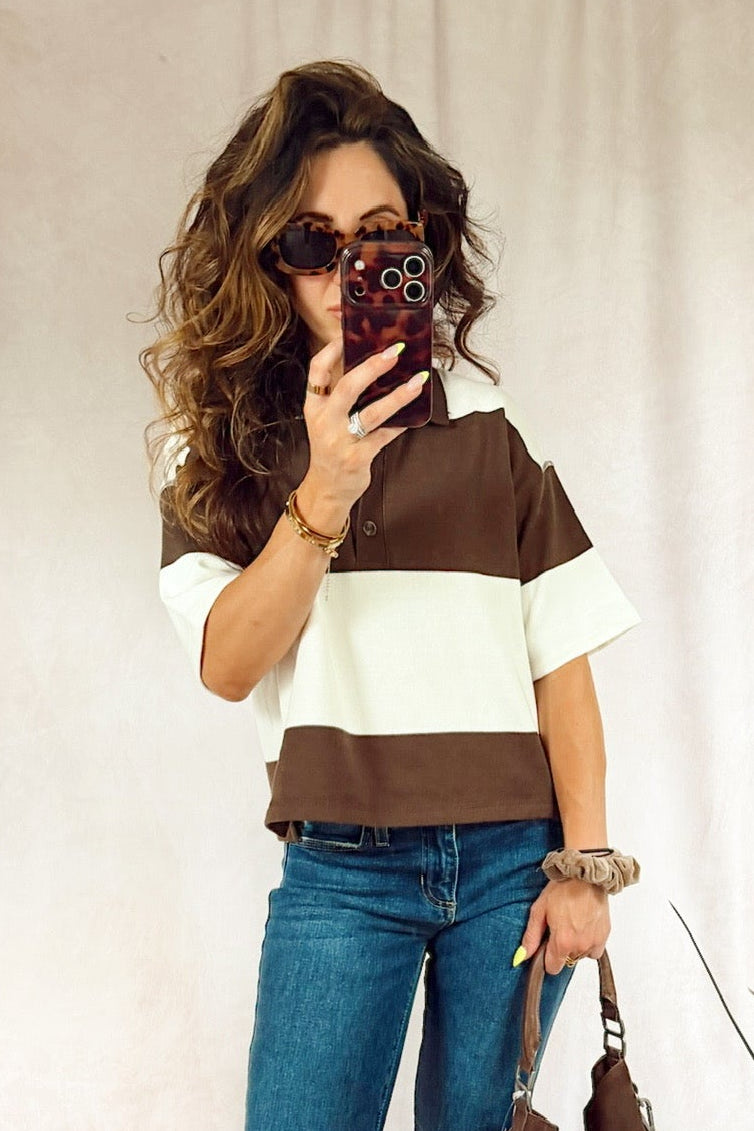 A woman takes a mirror selfie wearing a brown and white color-blocked short-sleeve top, blue jeans, and white sneakers, while holding a brown tote bag. She is standing in front of a white fabric backdrop with indoor plants visible on the sides.