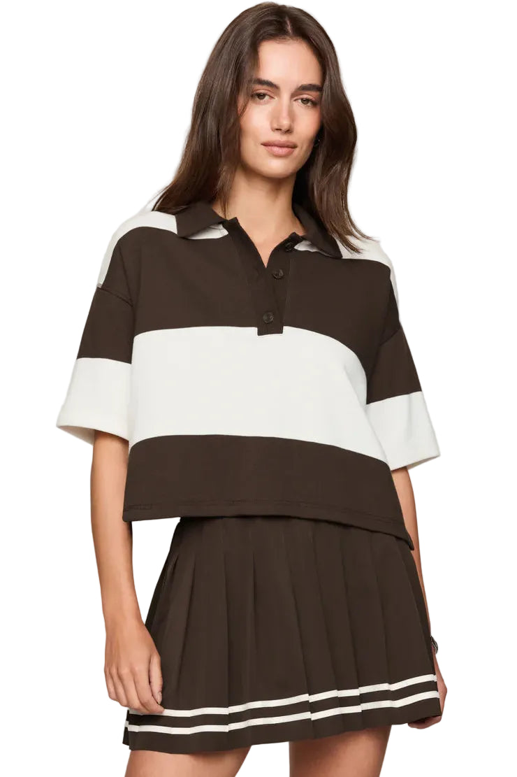 A woman modeling a color-blocked short-sleeve polo shirt and matching pleated mini skirt set. The set features dark brown and off-white horizontal stripes, with the skirt accented by double white stripes near the hem.