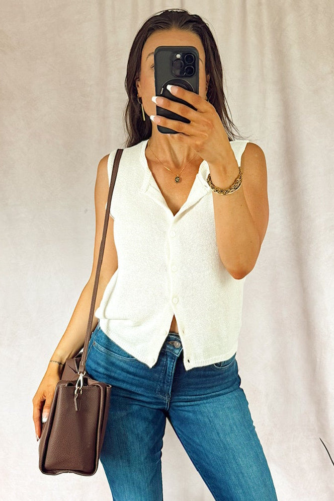 A woman takes a mirror selfie wearing a white sleeveless button-front top and blue flare jeans, holding a brown handbag. The photo is taken indoors in front of a light fabric backdrop with plants visible at the edges. z supply. good american.